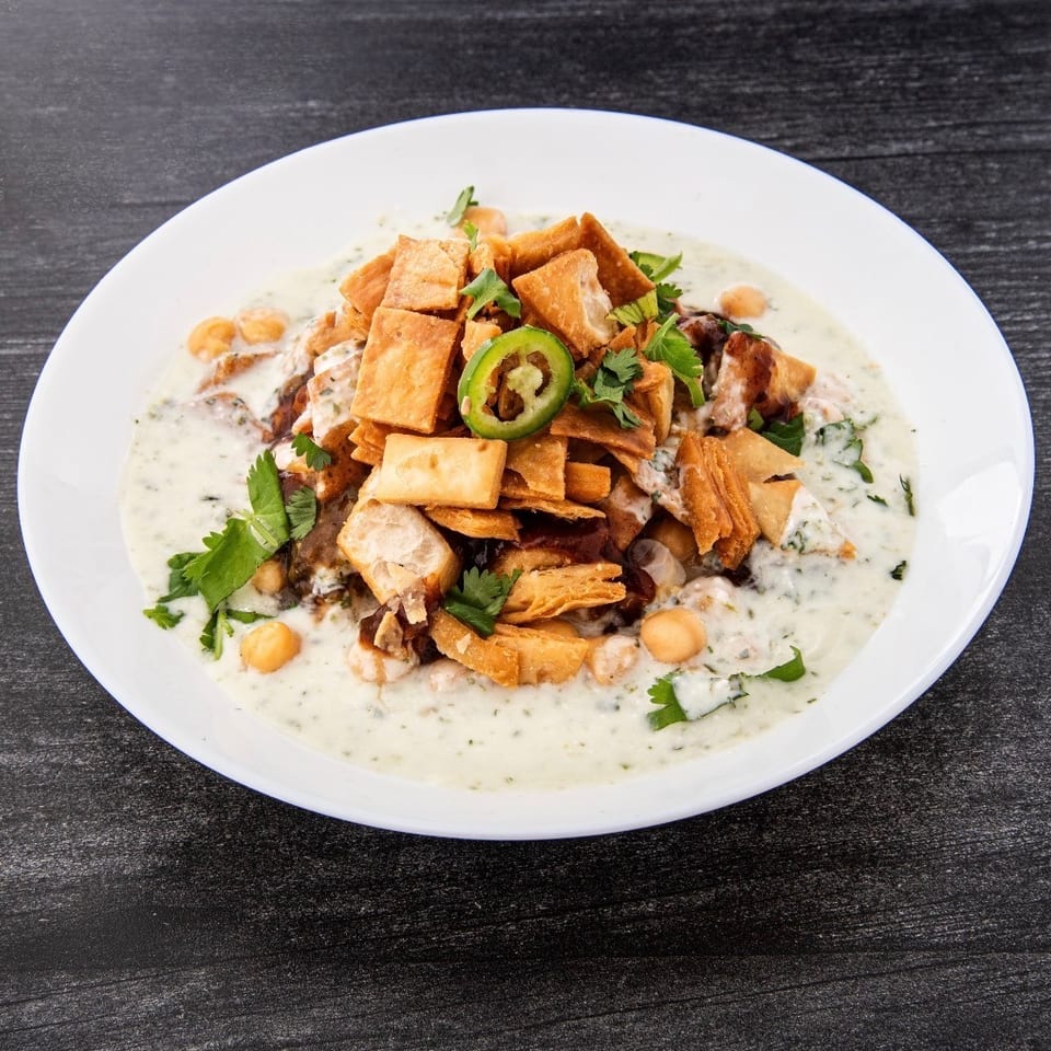 White plate of creamy chickpea stew topped with crispy pastry pieces, jalapeño slices, and fresh herbs on a dark wooden table.