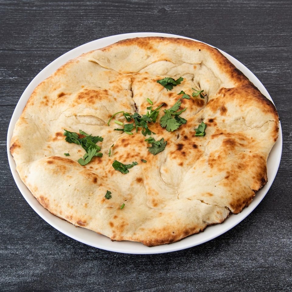 Large golden-brown flatbread garnished with fresh cilantro on a white plate atop a dark wooden table.