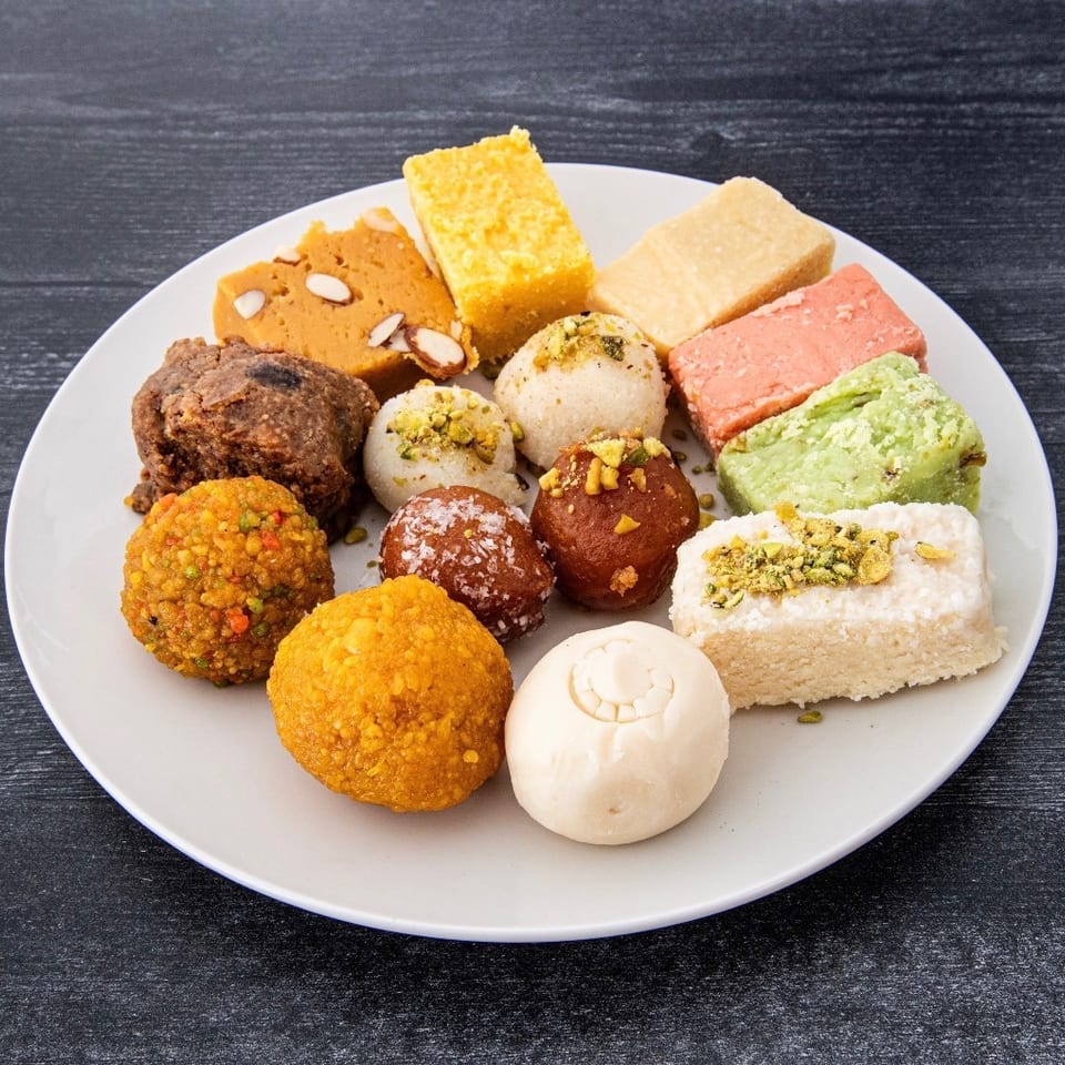 White plate filled with assorted colorful Indian sweets, including ladoos, fudge squares, and pistachio-topped confections.